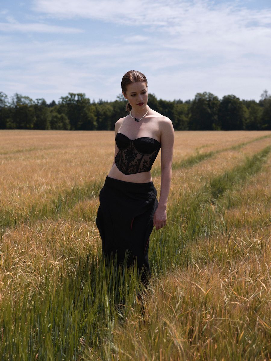 Eine junge Dame steht in einem Kornfeld. Sie trägt einen Pferdeschwanz und ein schwarzes, trägerloses Oberteil und einen schwarzen, engen Rock.