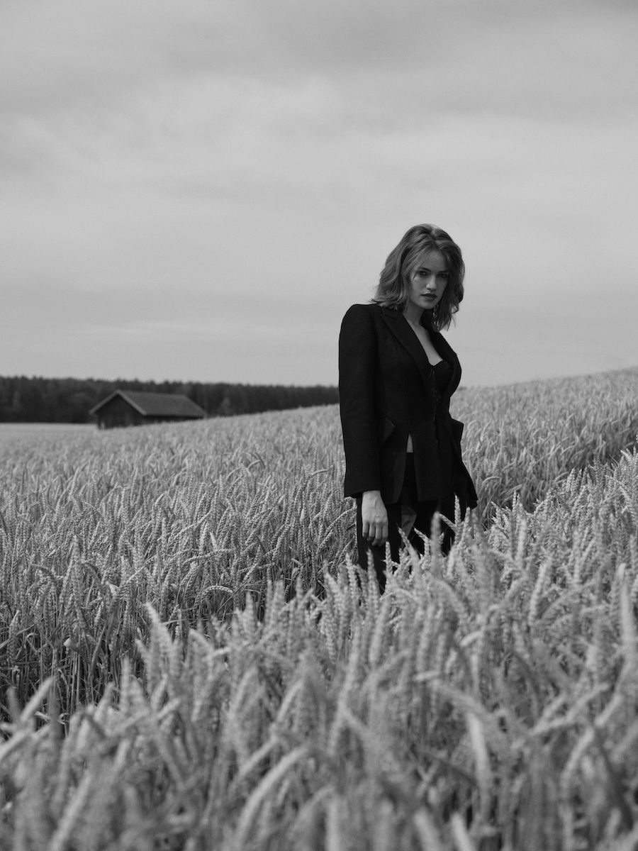Schwarz Weiß Foto einer jungen Dame, sie steht in einem Kornfeld. Sie trägt offenes, schulterlanges Haar und einen schwarzen Blazer, dazu eine schwarze Hose.