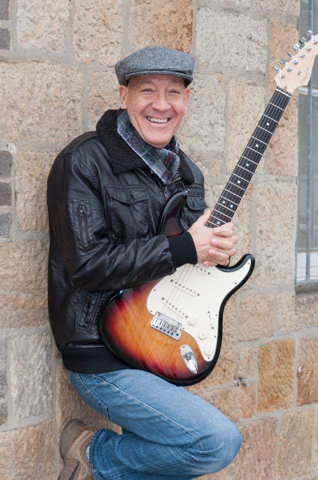 Ein Mann mit Mütze und Stratocaster in der Hand lächelt sympathisch in die Kamera.