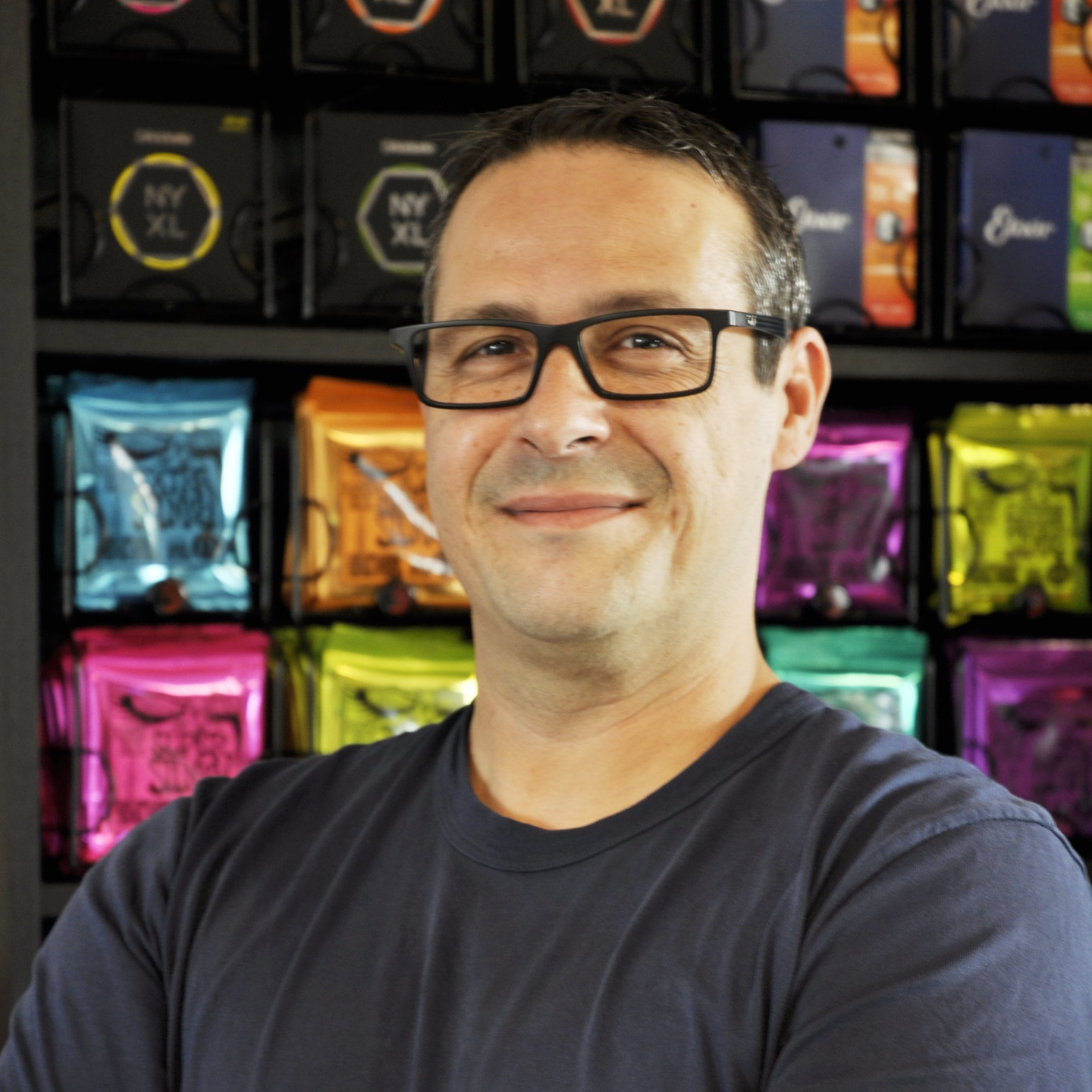 Ein Mann mit Brille lächelt freundlich in die Kamera. Hinter ihm ist eine Wand mit Gitarrensaiten.