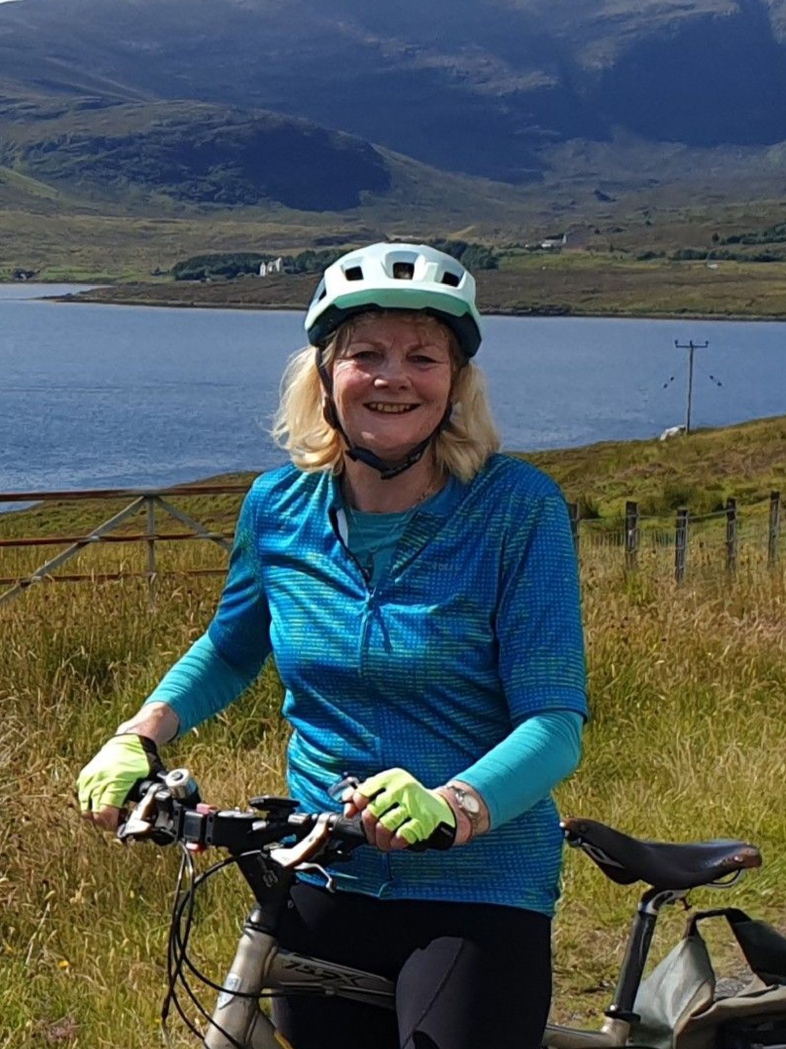 Woman (felicity) in blue cycling top and helmet holding a hybrid bike in front of a lake. Possibly peak district.