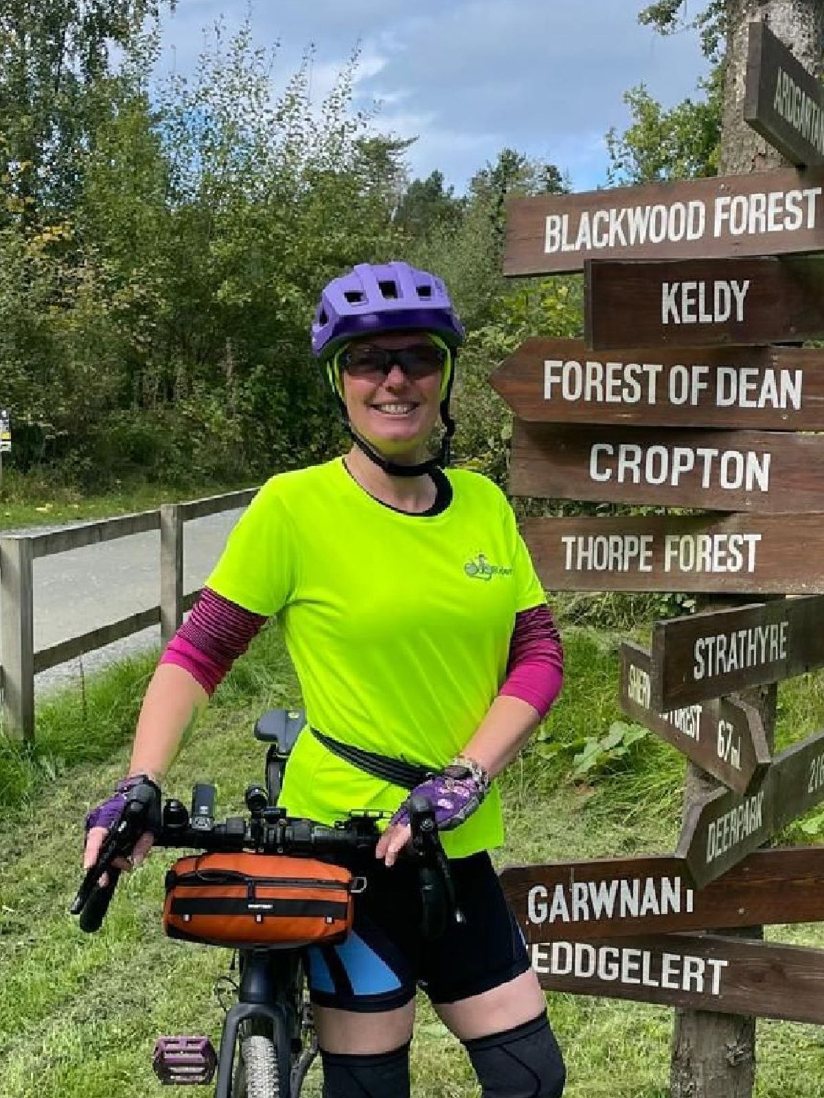 Woman (Helen) in lime green t-shirt and sunglasses holding touring bike next to a sign with lots of directions on it.