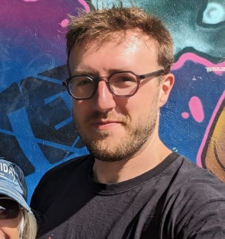 Head and shoulders shot of a white man wearing square glasses stood in front of wall of graffiti art.