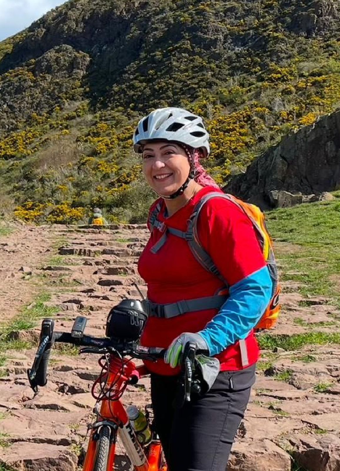 Woman (Samina) with touring bike and red and blue top and helmet against background of a hill. Possibly peak district.
