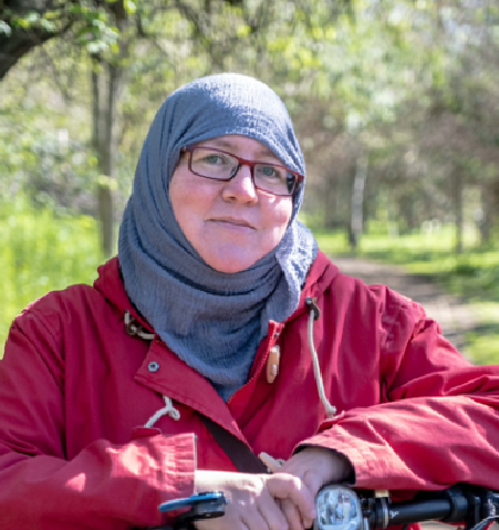 White woman wearing square, red glasses and a blue headscarf. She is leaning over the handlebars of a bike. Background is a park.