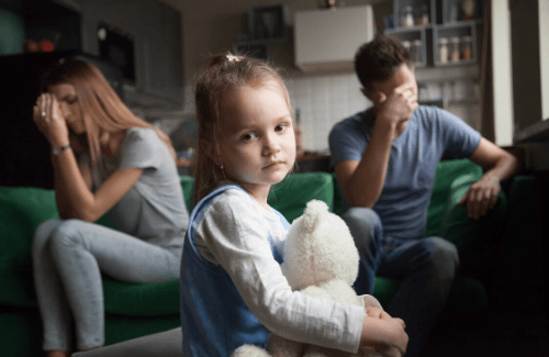 Niña sujetando peluche triste y sus padres enfadados por detrás