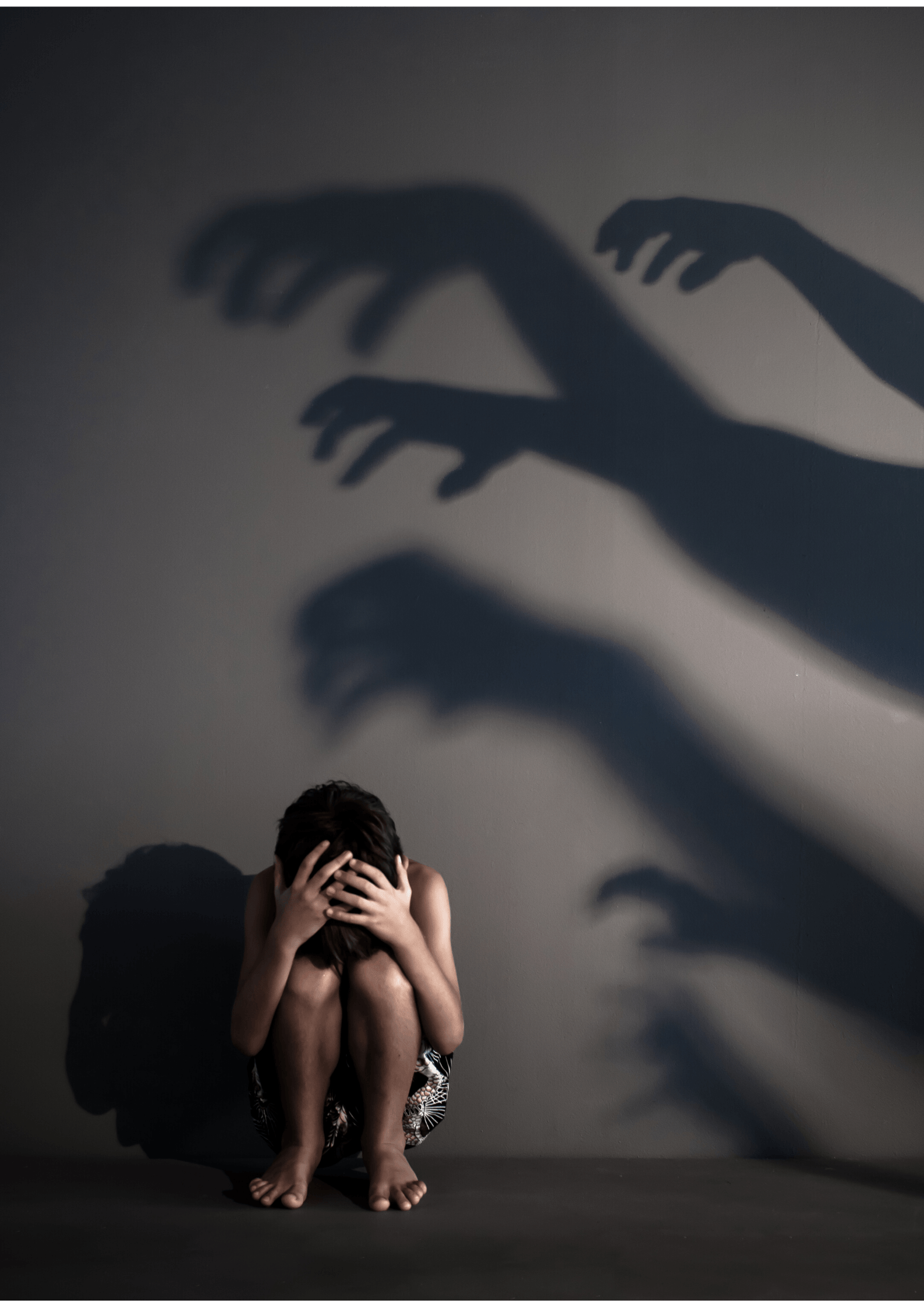 Niño sentado en el suelo enterrando la cabeza entre las rodillas asustado con sombras de manos que intentan agarrarle