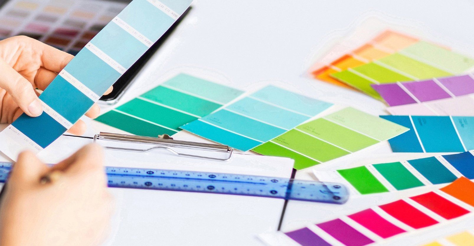 Auf dem Bild ist eine Nahaufnahme zu sehen, die eine Szene aus einem kreativen oder gestalterischen Arbeitsumfeld zeigt. Im Vordergrund halten zwei Hände eine Farbpalette mit verschiedenen Abstufungen von Blau- und Türkistönen.