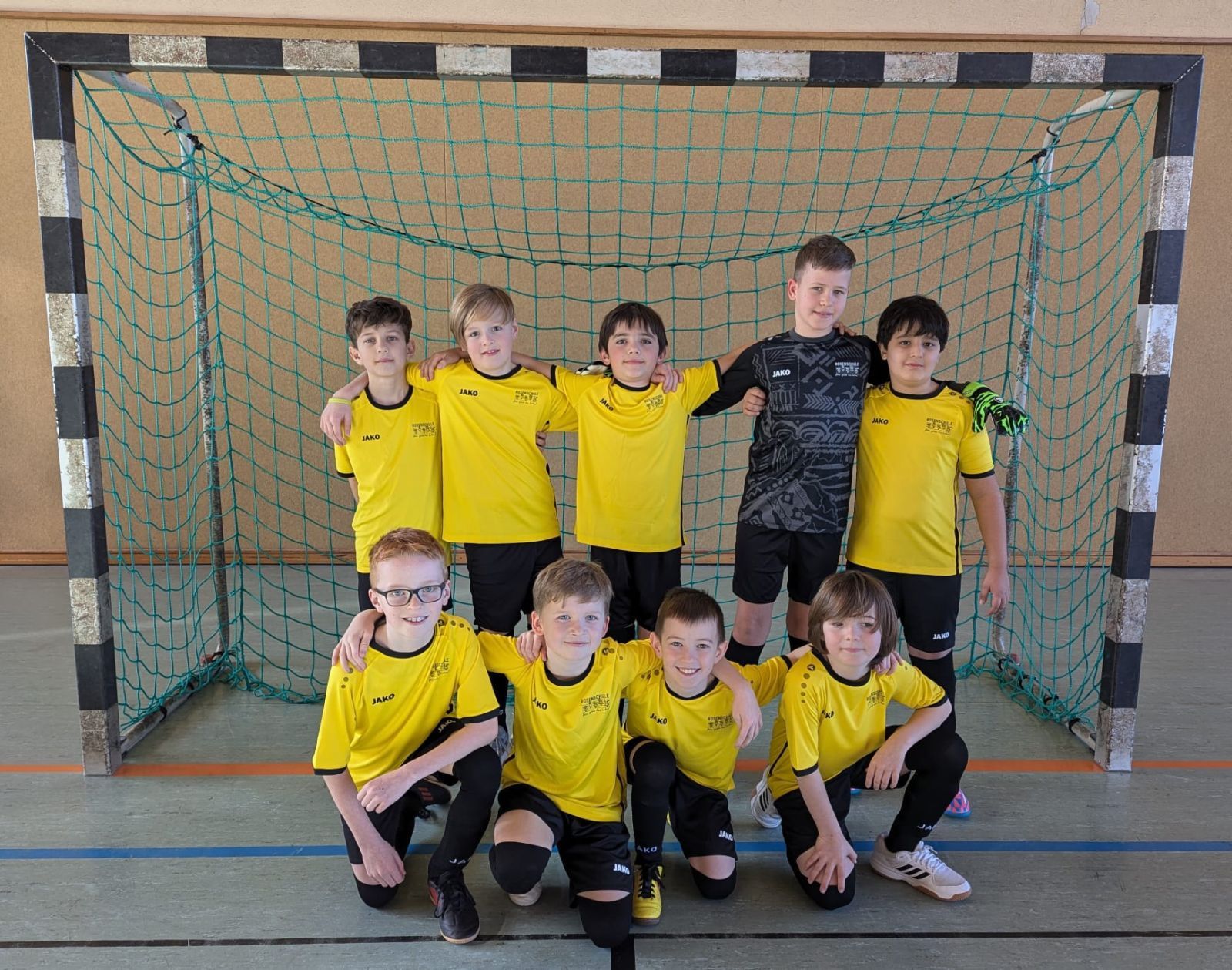 Mannschaftsbild des Rosenschul-Teams beim Grundschul-Cup Mannschaftsbild Fußballmannschaft der Rosenschule. 9 Schüler der Schulmannschaft stehen in schwarz-gelben Fußballtrikots in einem Hallenfußballtor