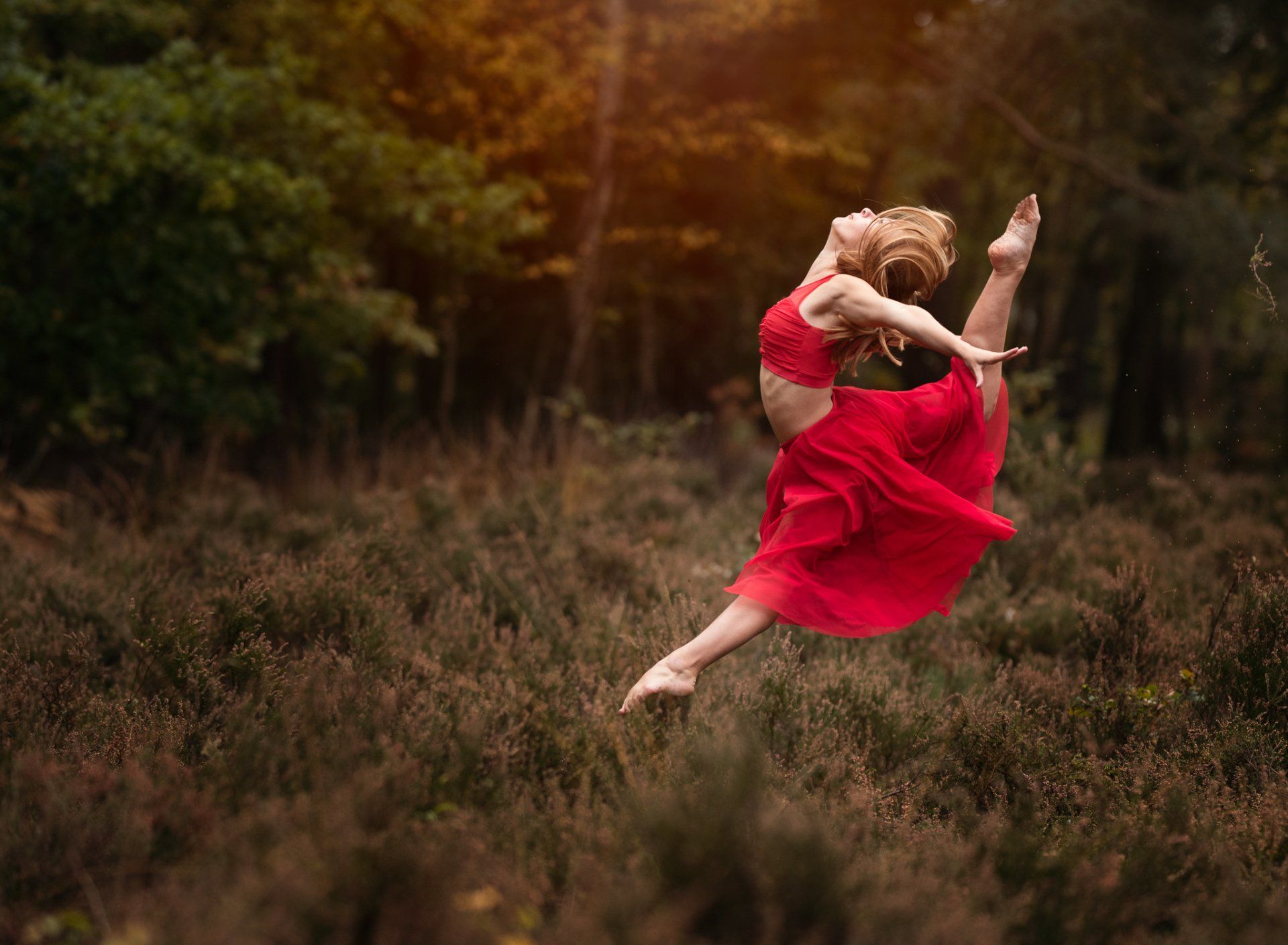Dancer dressed in red performing a leap in heather field with sun setting  behind her