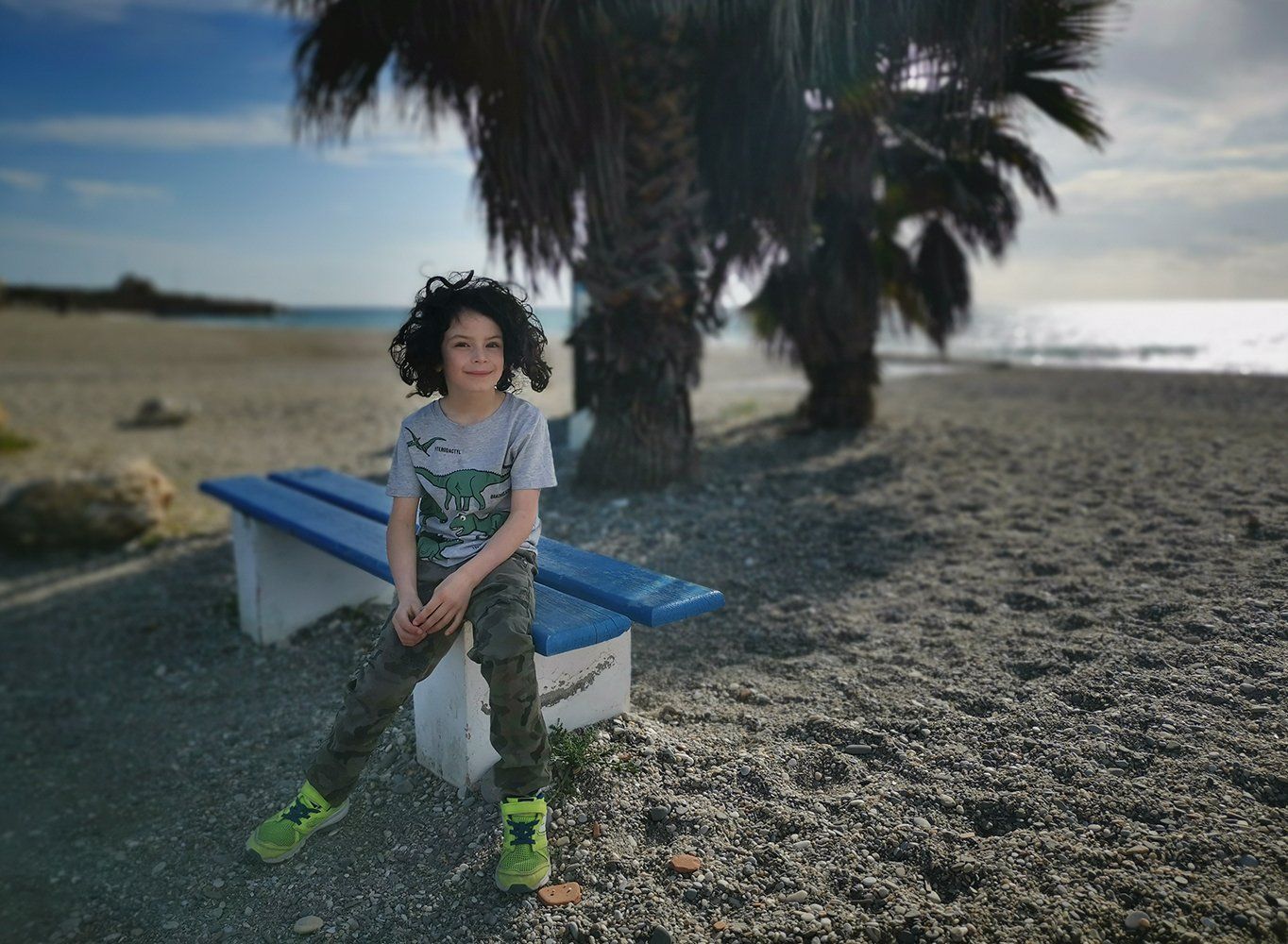 Beautiful boy with curly brown hair sitting on bench under a palm tree
