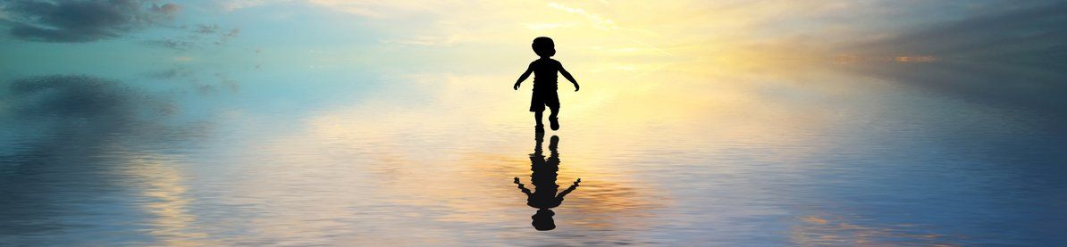 Silhouette of a child with a sunset behind them, and their shadow reflected on calm water.