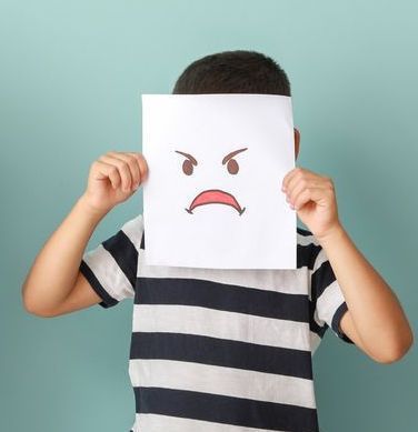 A child in a stripy t-shirt. He is holding a piece of paper over his face. On the paper is a drawing of a grumpy face.