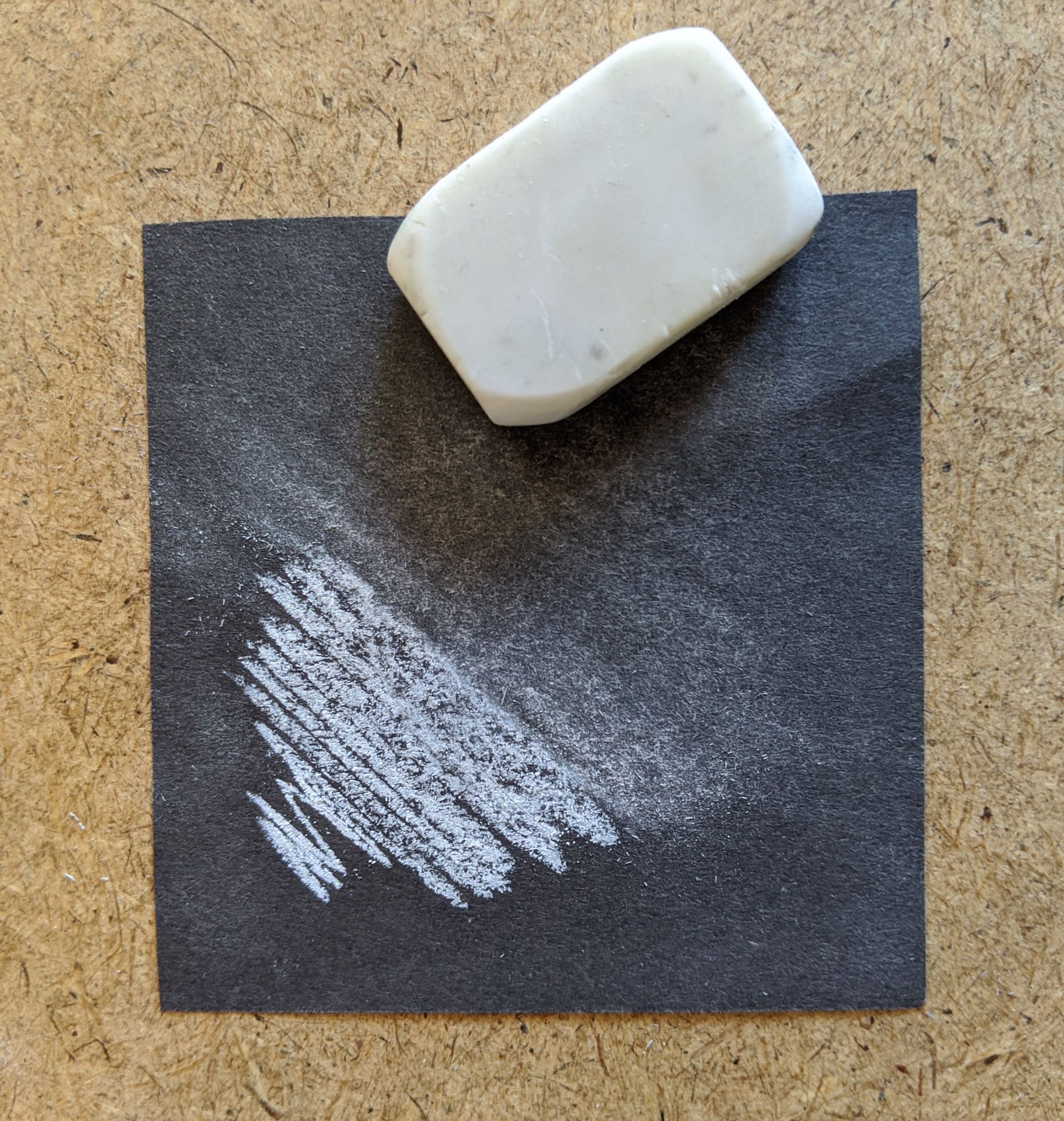 A dark coloured piece of paper with some white scribble on it. Half of it has been rubbed out with the eraser.