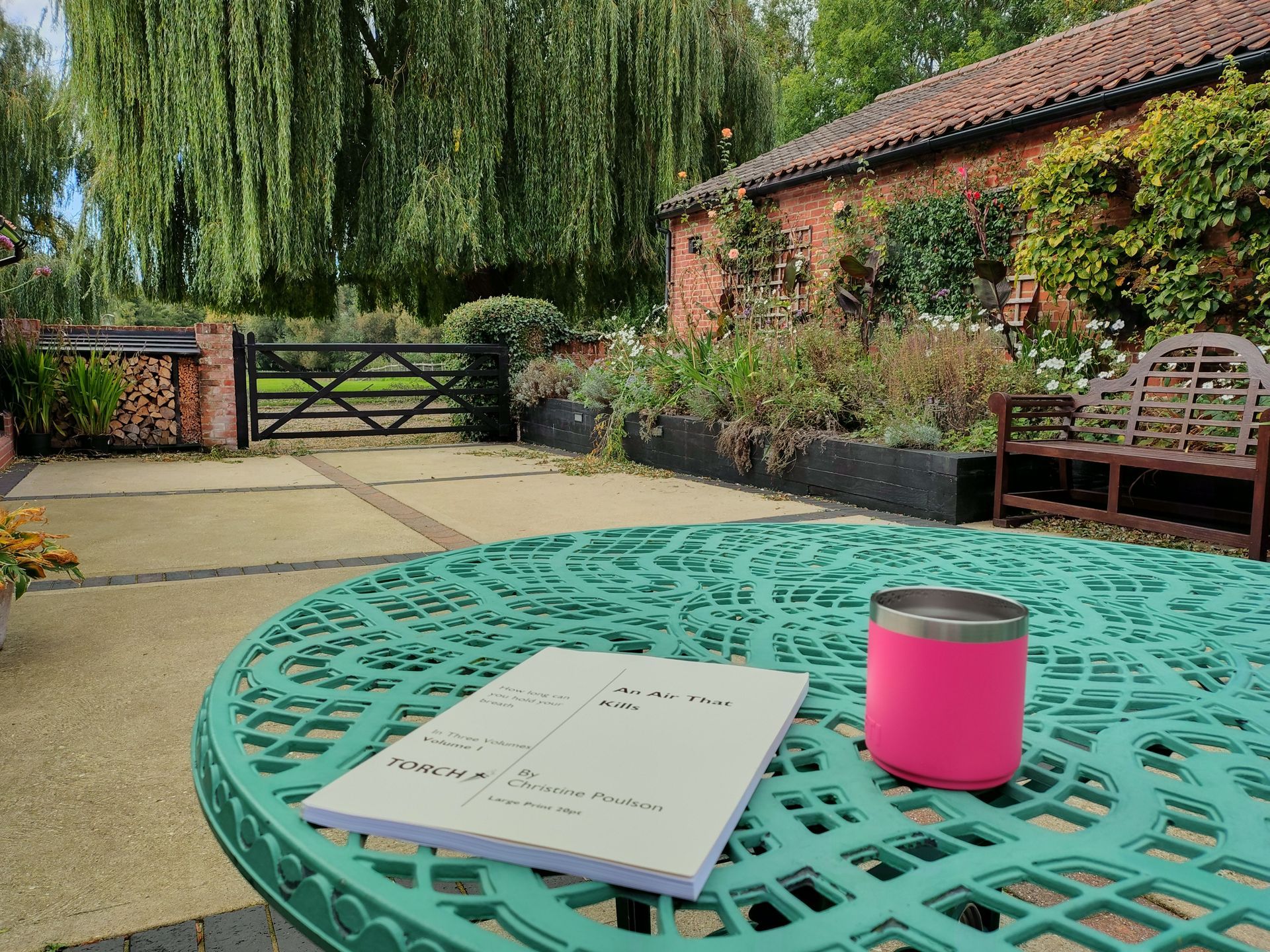 A photo taken in a beautiful courtyard. In the foreground is a light green iron table with patterned cutouts. On top of it is a pink thermos cup and a large print novel from torch trust. IN the distance is a wooden gate and beyond that a very tall willow tree