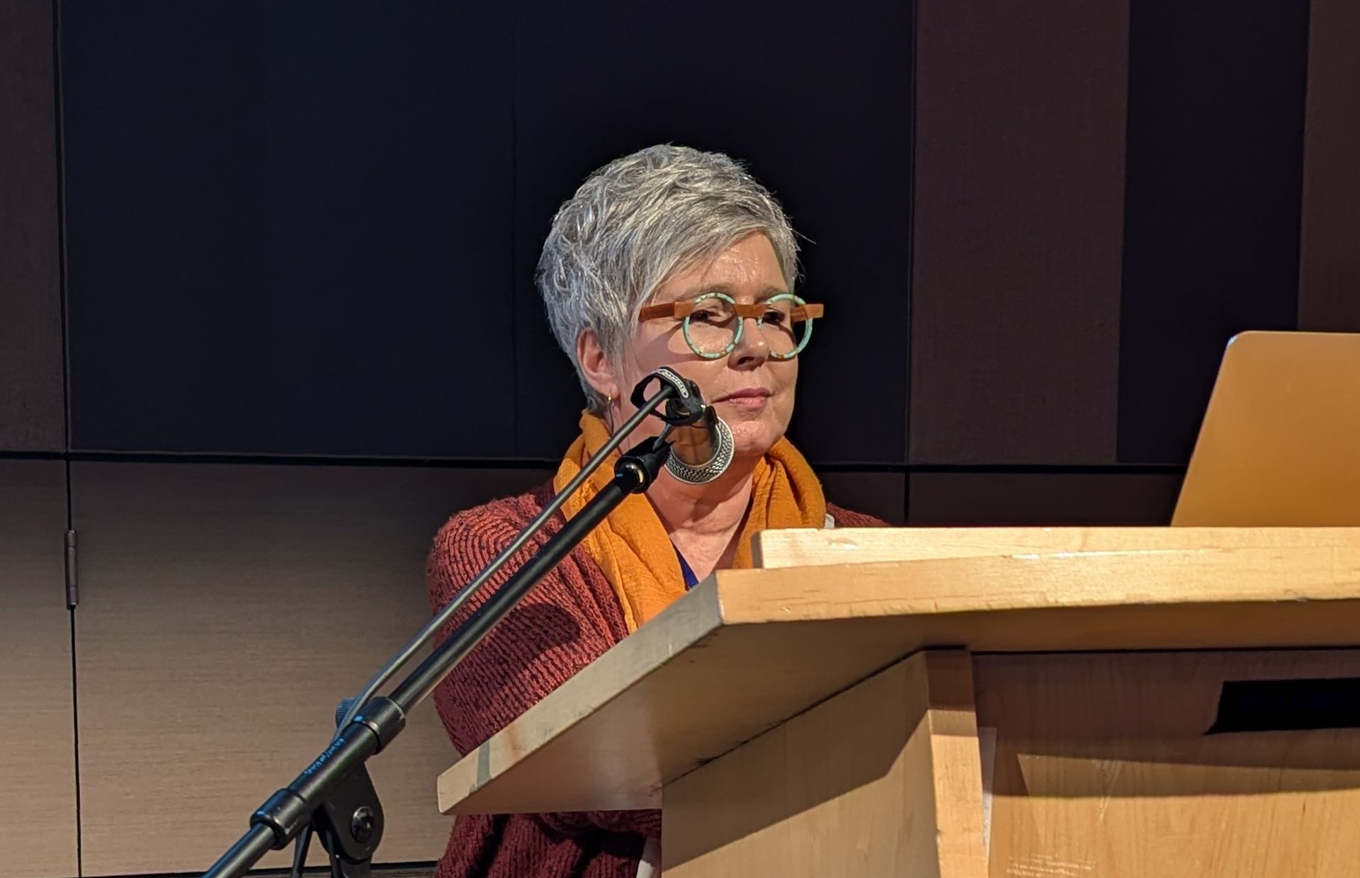 Picture of Kay behind a lectern that is too high for her. There is a microphone to her right.