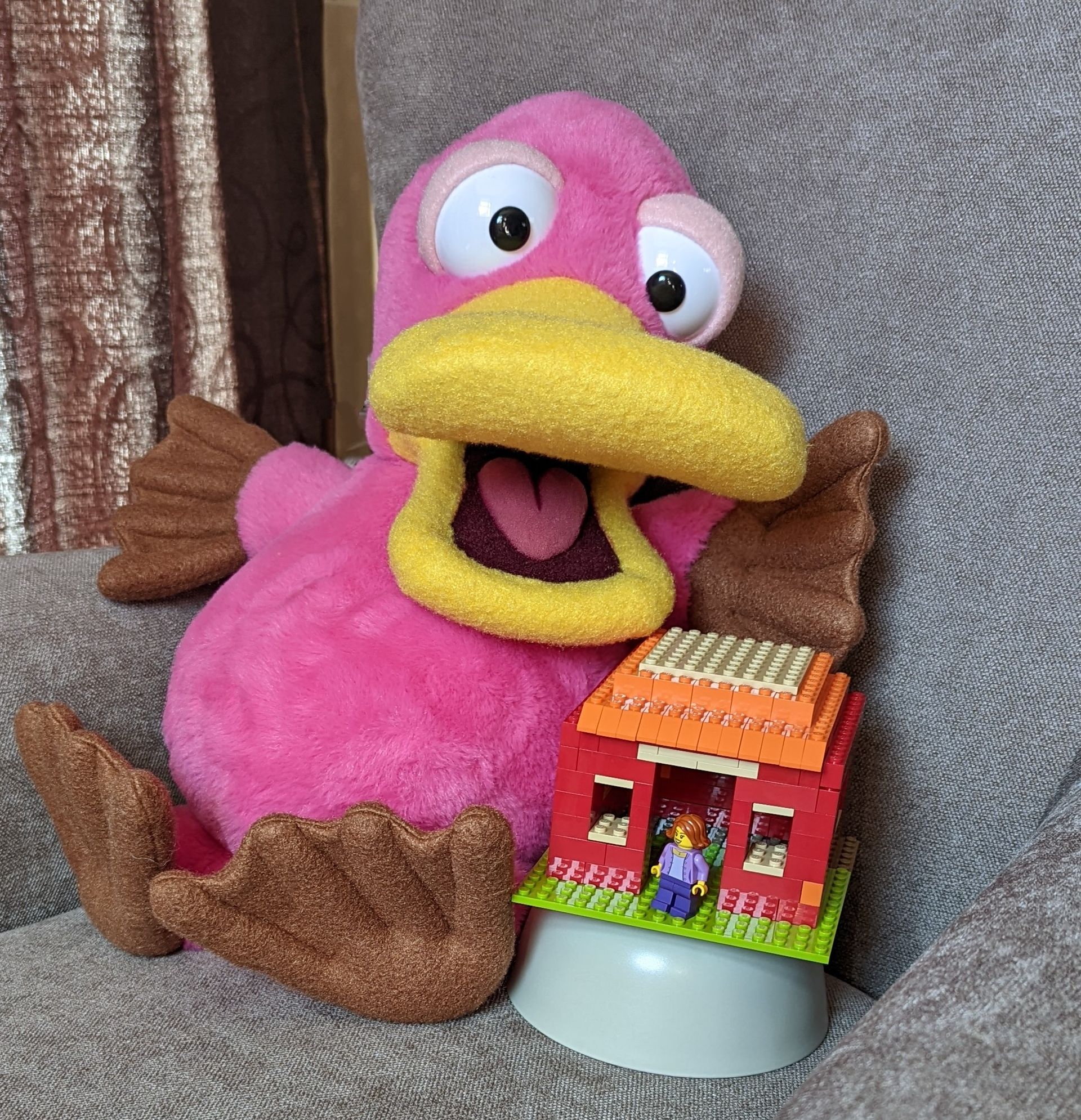 Theo, the pink platypus puppet. He has a small lego built house next to him. The house has a lego person in the doorway.