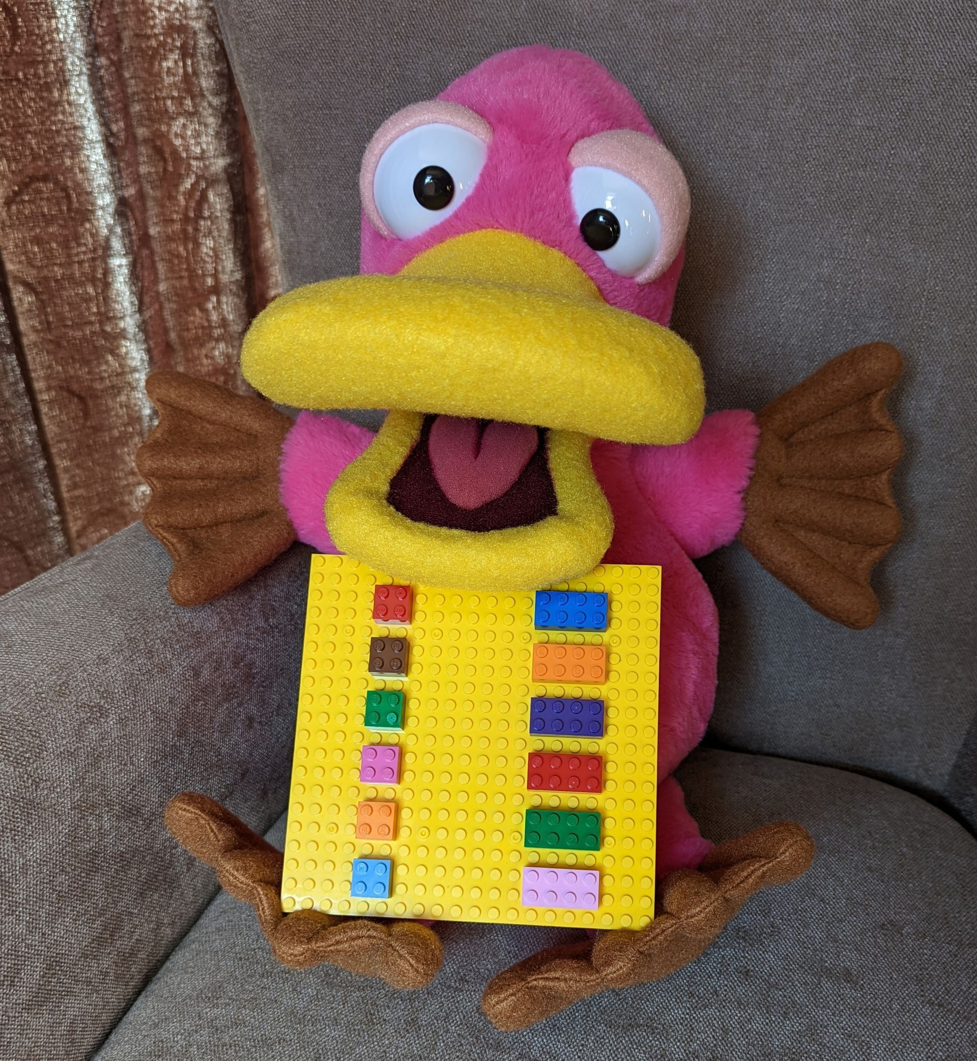 Theo the pink platypus puppet holding a lego base board. There is a downward line of different coloured lego on each side.