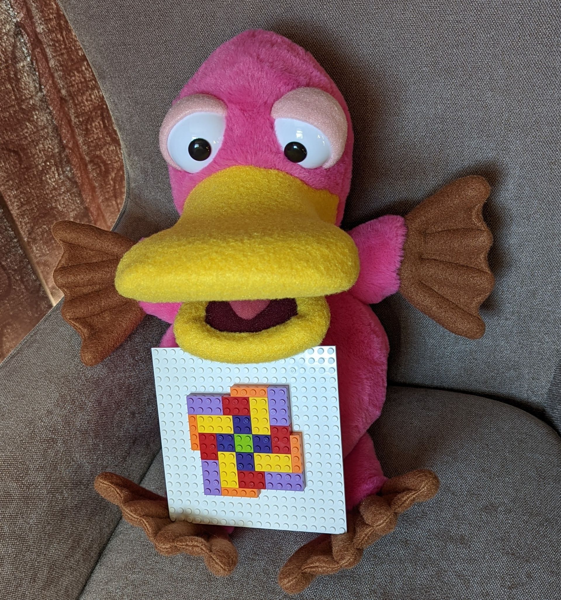 Theo the pink platypus puppet. He is holding a Lego base board with a mosaic pattern built with different coloured bricks on it.