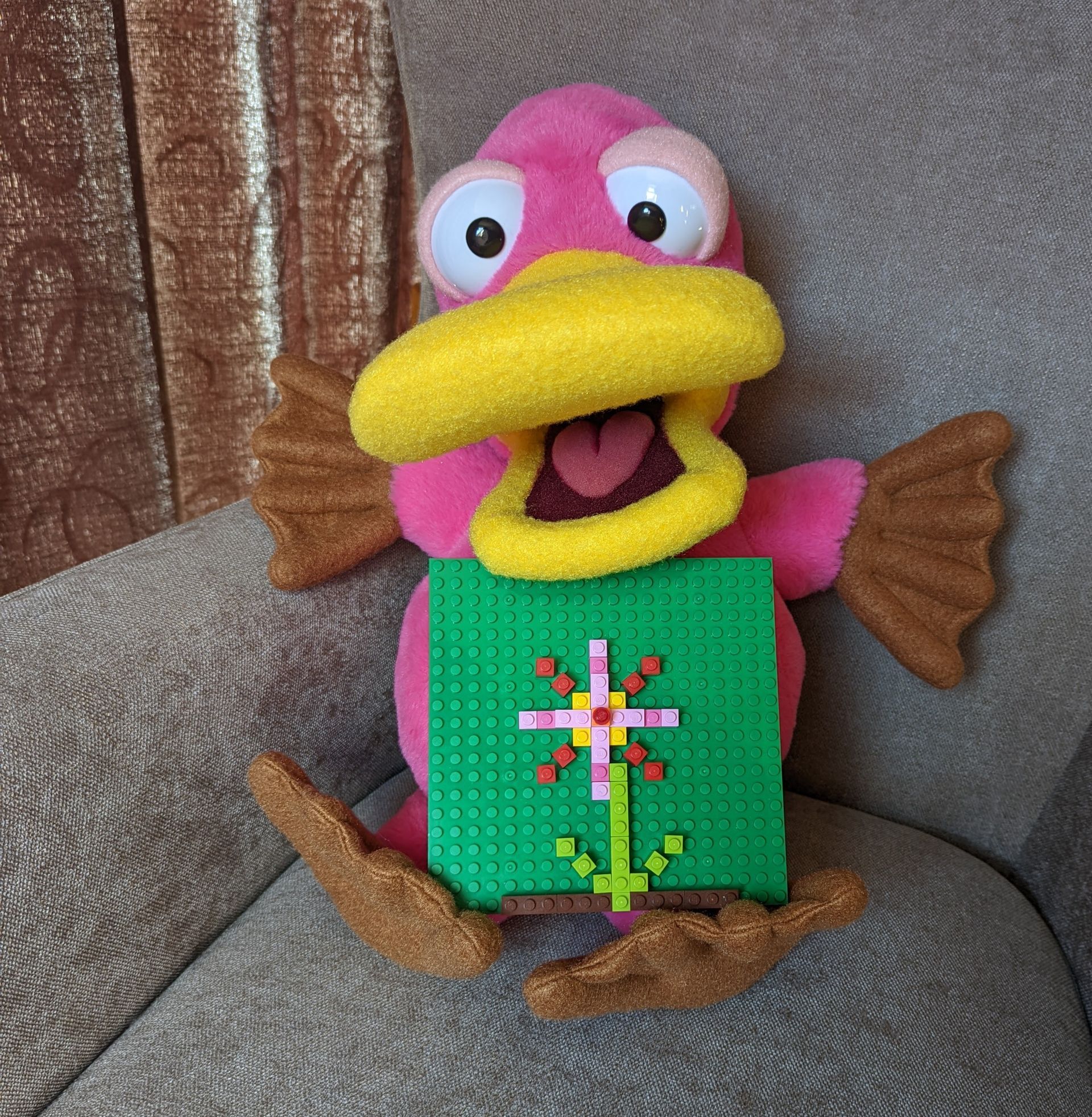 Theo the pink platypus puppet. He is holding a lego baseboard with a lego built flat flower picture on it.