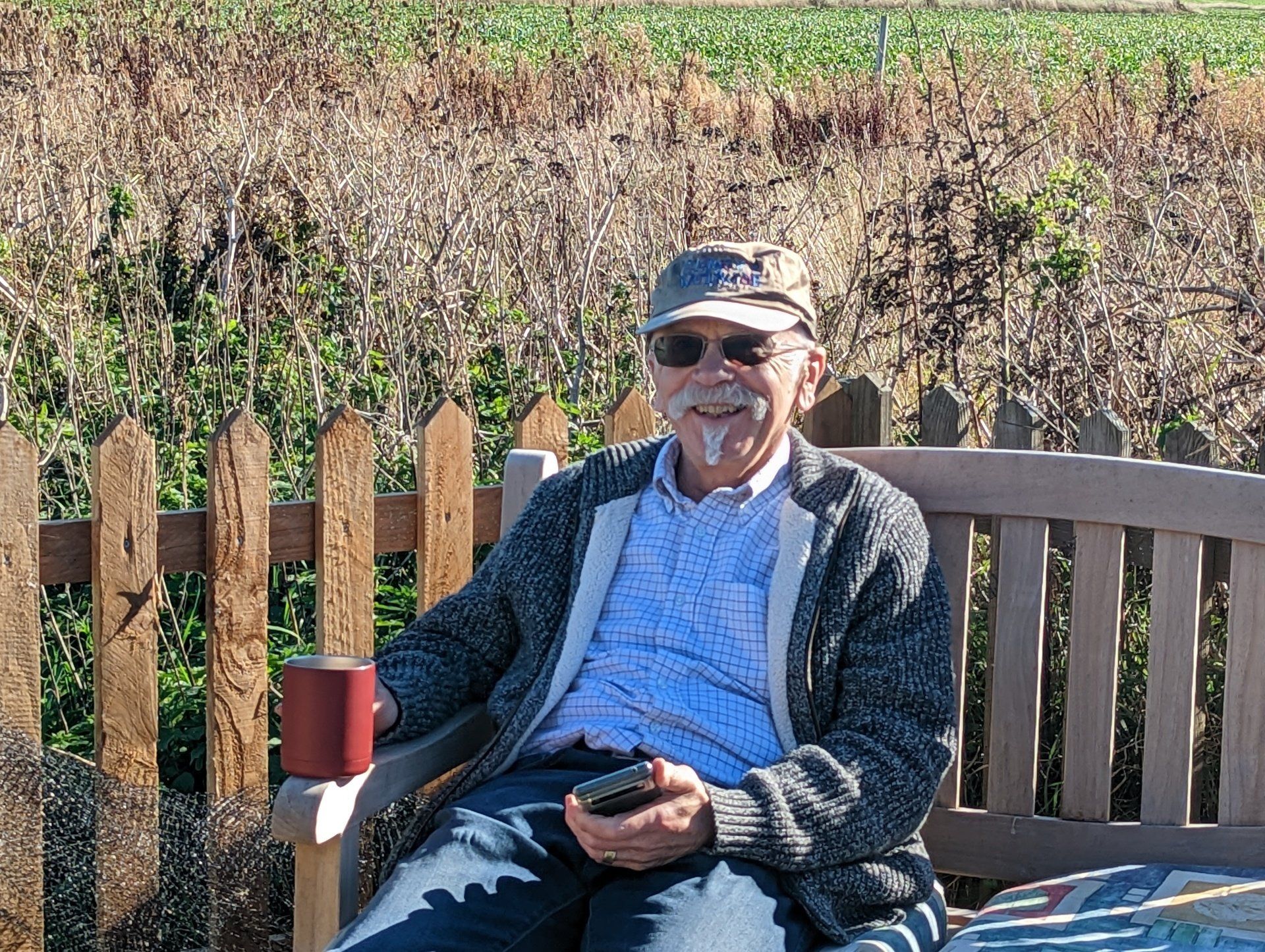 Steve sitting on a wooden bench enjoying the sunshine on holiday. He has a mug of tea in his hand