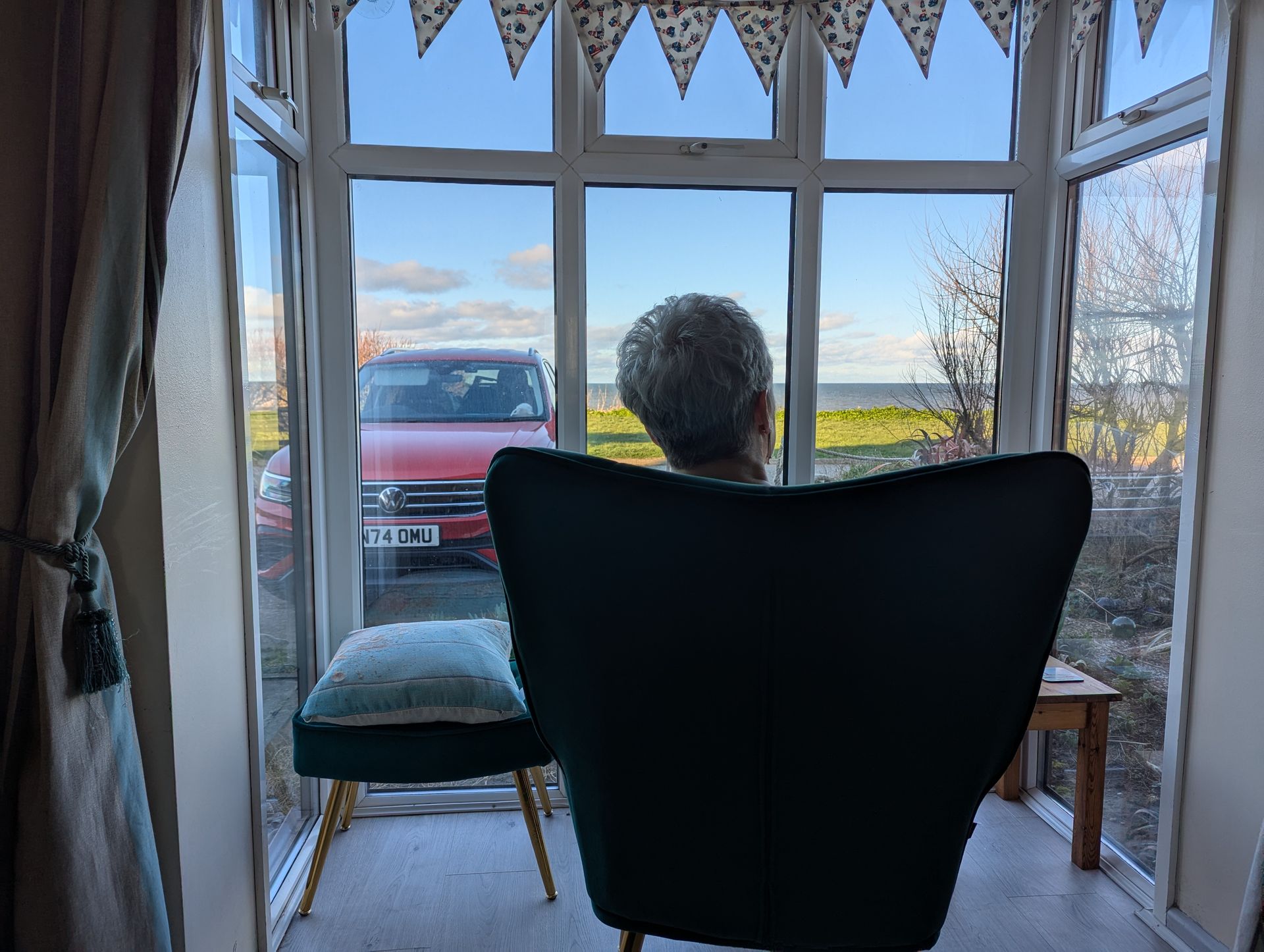 Kay is sitting in a chair looking out of the window. Beyond the window is a grassy area and beyond that is the sea.