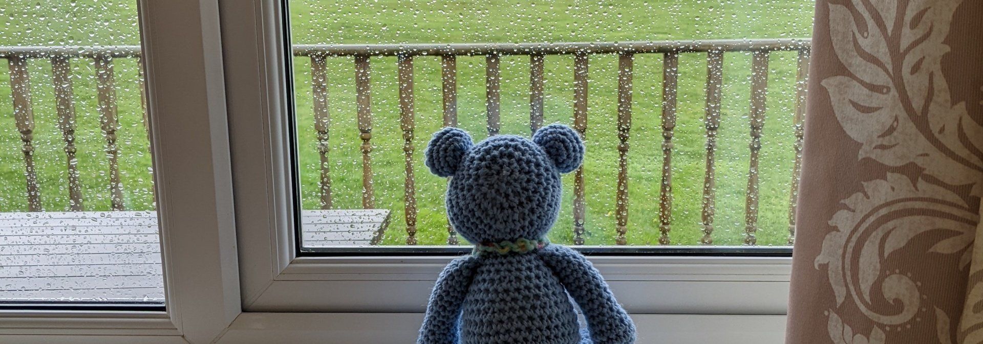 Photo of a blue crocheted bear looking out of a rain soaked window