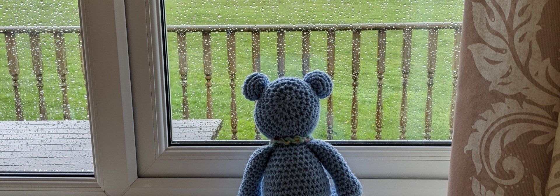 Photo of a blue crocheted bear looking out of a rain soaked window.
