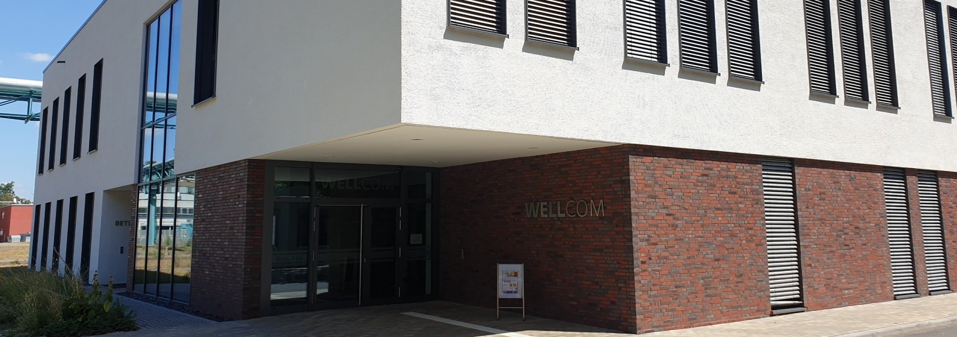 Moderner zweigeschossiger Gebäudeeingang mit großen Fensterflächen und Beleuchtung – vermutlich der Eingang des Gesundheitszentrums.