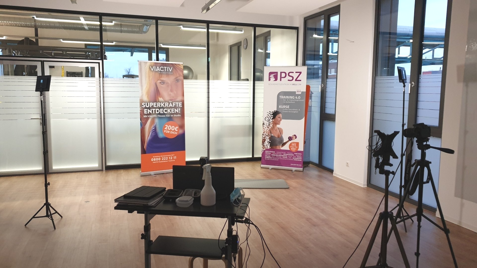 Bright training room with wooden floor, two roll-up banners, a table with a laptop, and camera equipment on tripods.