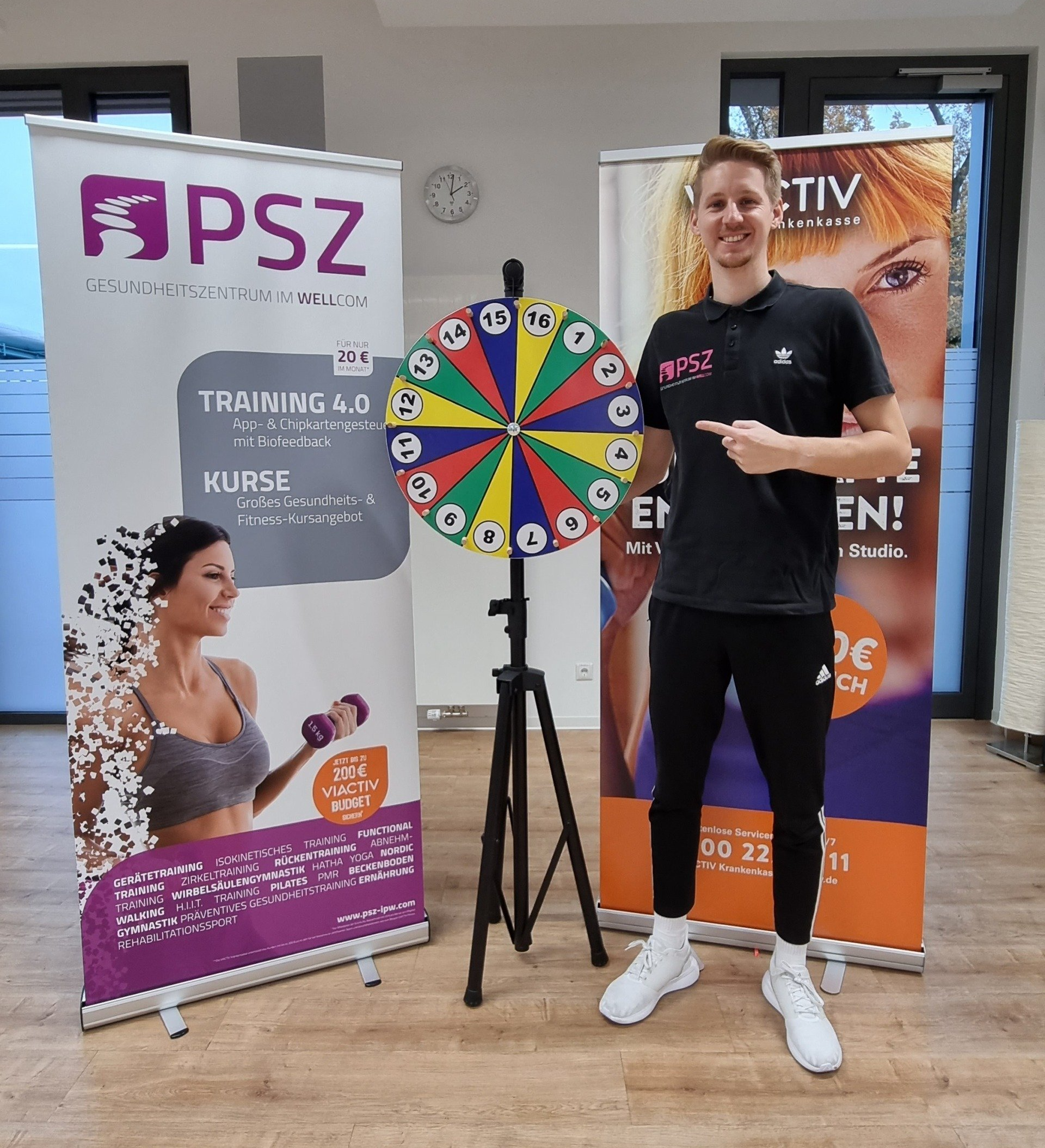 Junger Mann im PSZ-Shirt steht lächelnd neben einem bunten Glücksrad, zwischen zwei Werbebannern für das PSZ-Gesundheitszentrum.