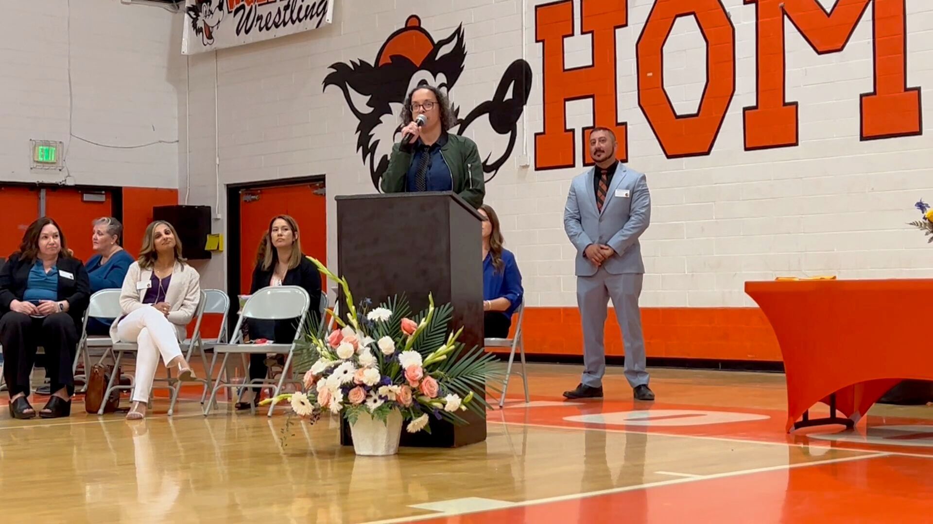 Me standing in the Woodland High School gym in June 2024, announcing the first David Breaux Memorial Scholarship Award, orange, black, and white Woody the Wolf mascot and white walls in the background