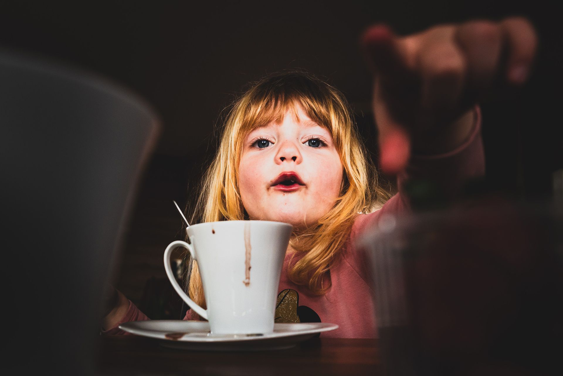 mädchen sitzt vor weißer tasse am tisch und zeigt in richtung des fotografen