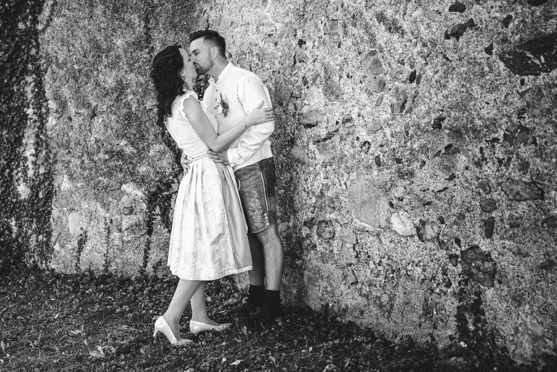 Ein Hochzeitspaar steht vor einer Mauer in Chieming und küsst sich. Die Situation sieht ungestellt und zufällig aus, sie ist aber bewusst inszeniert, damit es gut aussieht.
