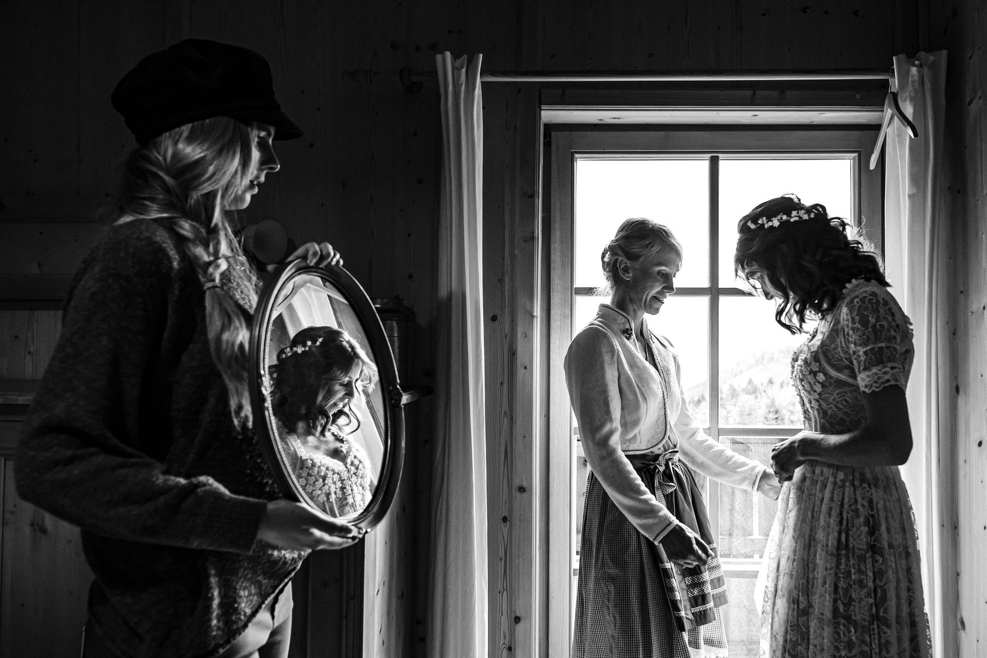 hochzeitsfotograf schliersee almbad huberspitz braut und schwester stehen im fensterlicht dritte person hält spiegel