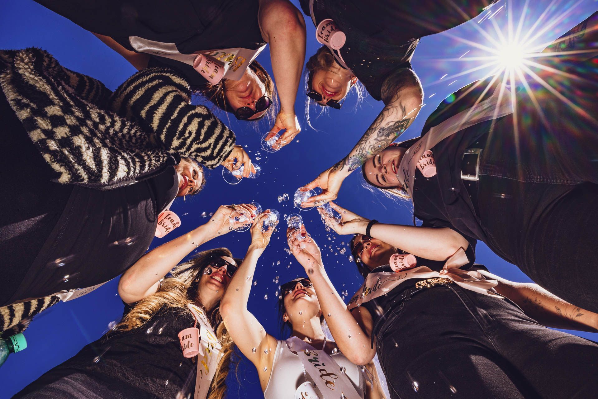Dynamisches JGA Fotoshooting: Coole Braut-Crew mit Sonnenbrillen stößt aus der Froschperspektive vor strahlend blauem Himmel an, Getränke spritzen in die Luft