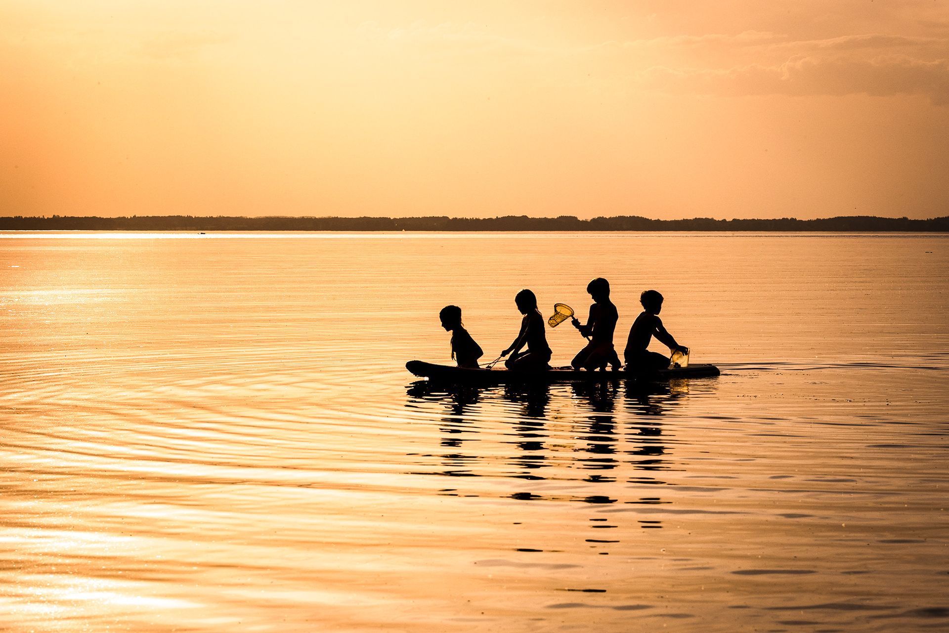 4 Kinder als Silhouetten sitzen im Sonnenuntergang auf einem SUP auf dem Chiemsee 4 Kinder als silhouette sitzen auf SUP im sonnenuntergang auf dem chiemsee