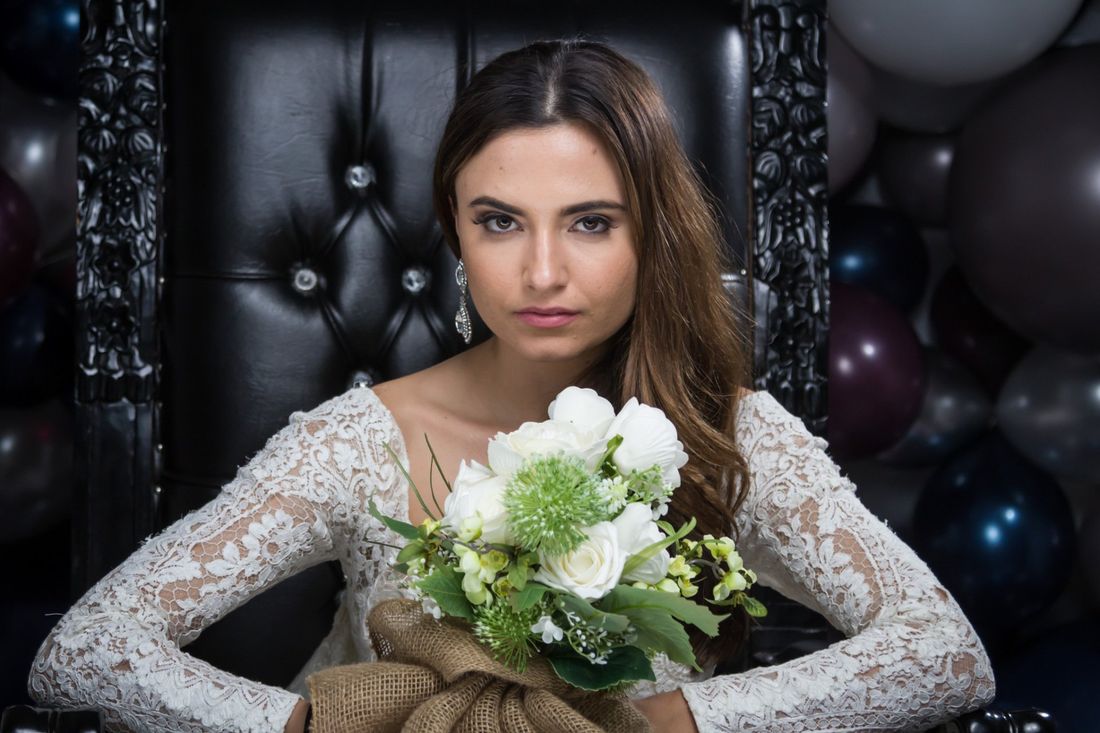 An indoor portrait of a woman in a white dress holding a bouquet of flowers sitting in a black leather chair
