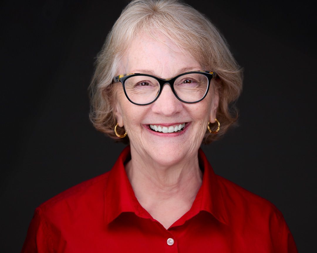 A woman with blond hair and glasses wearing a red button up shirt against a dark background.