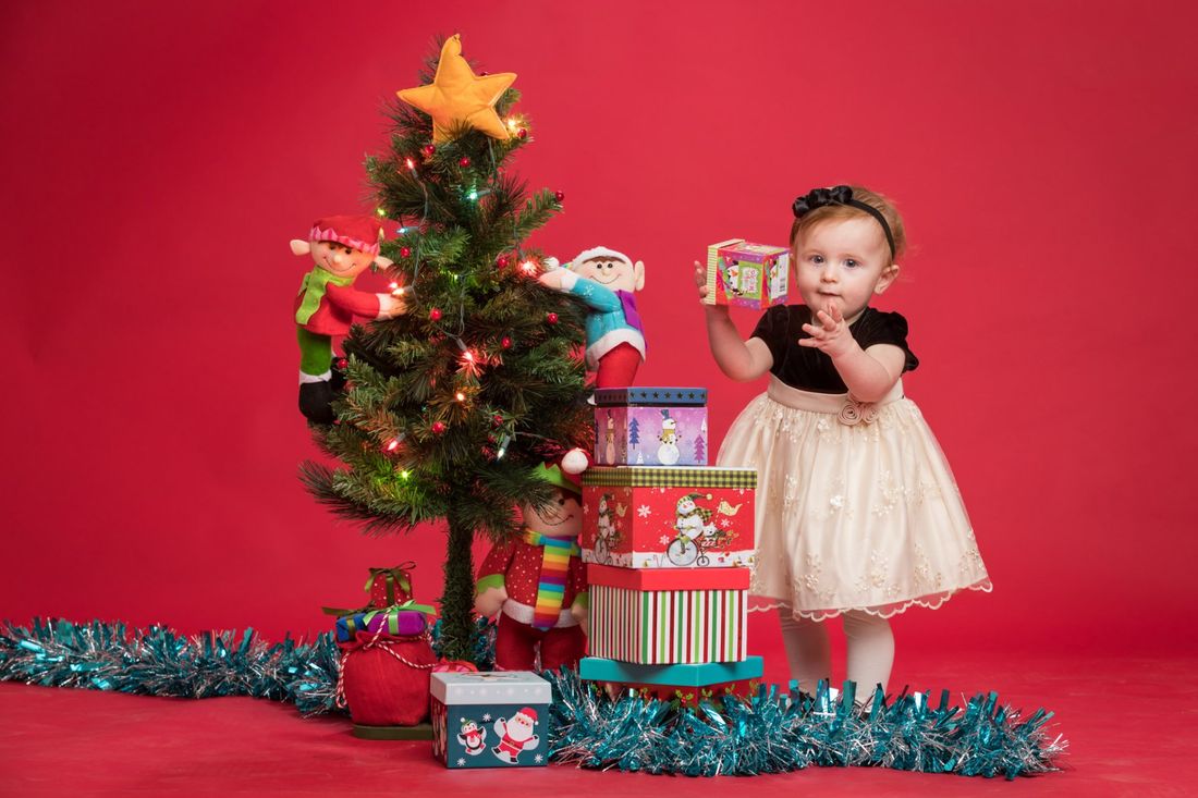 A studio portrait of a small girl wearing a black dress with white tulle standing next to a Christmas tree with presents with a red background