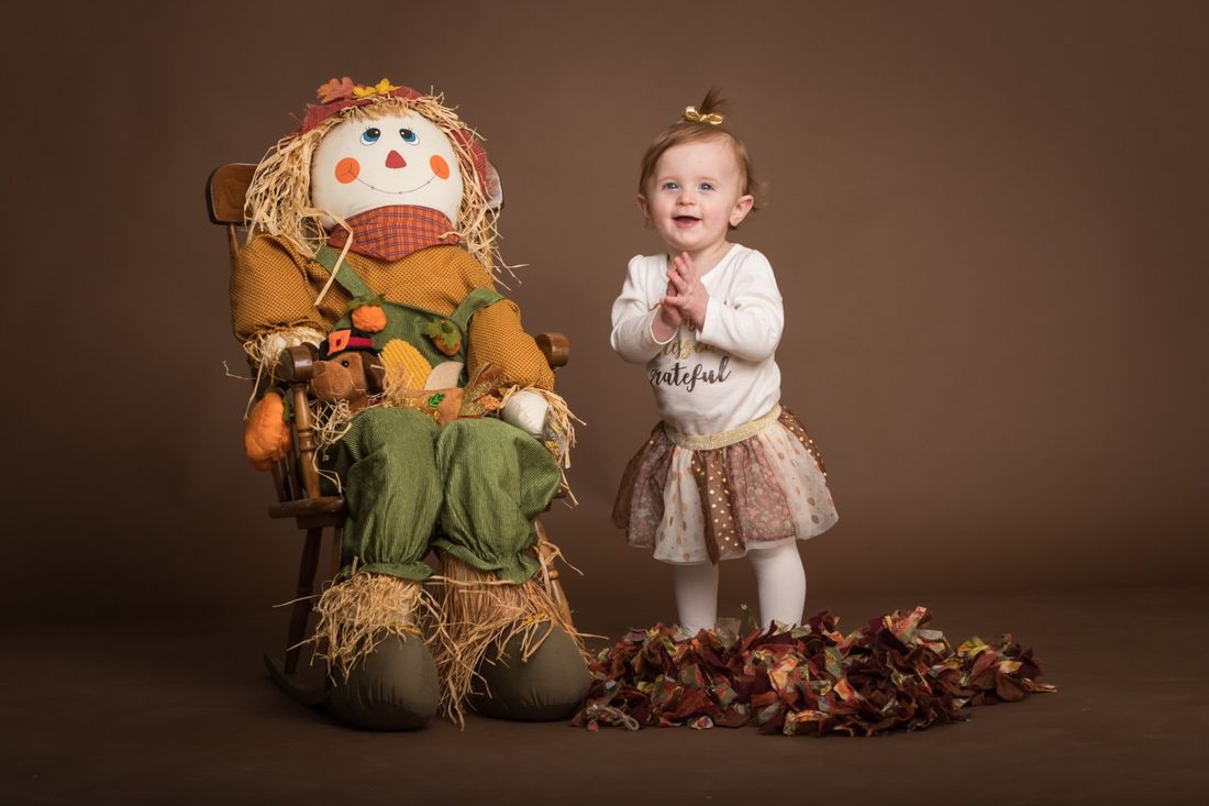 A studio portrait of a small girl standing next to a stuffed scarecrow for fall portraits with a brown background