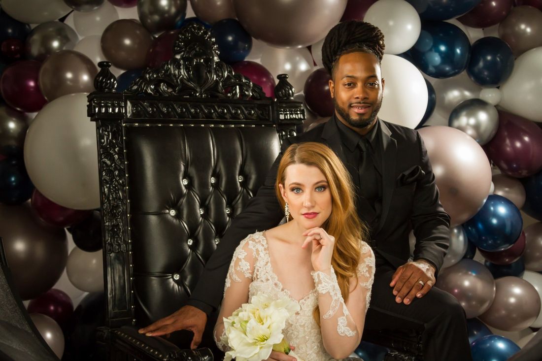 A styled portrait of a couple on a black leather chair with black, white and grey balloons in the background
