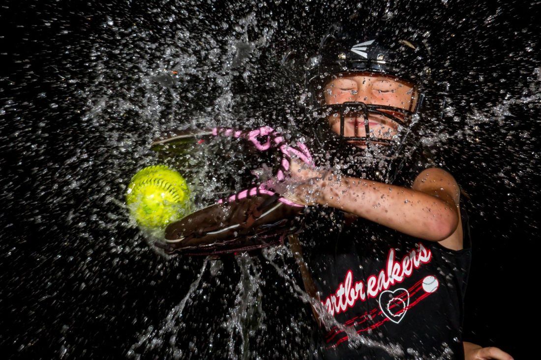 A creative portrait of a softball catcher catching a softball in her glove as water exploded from the impact against a black background