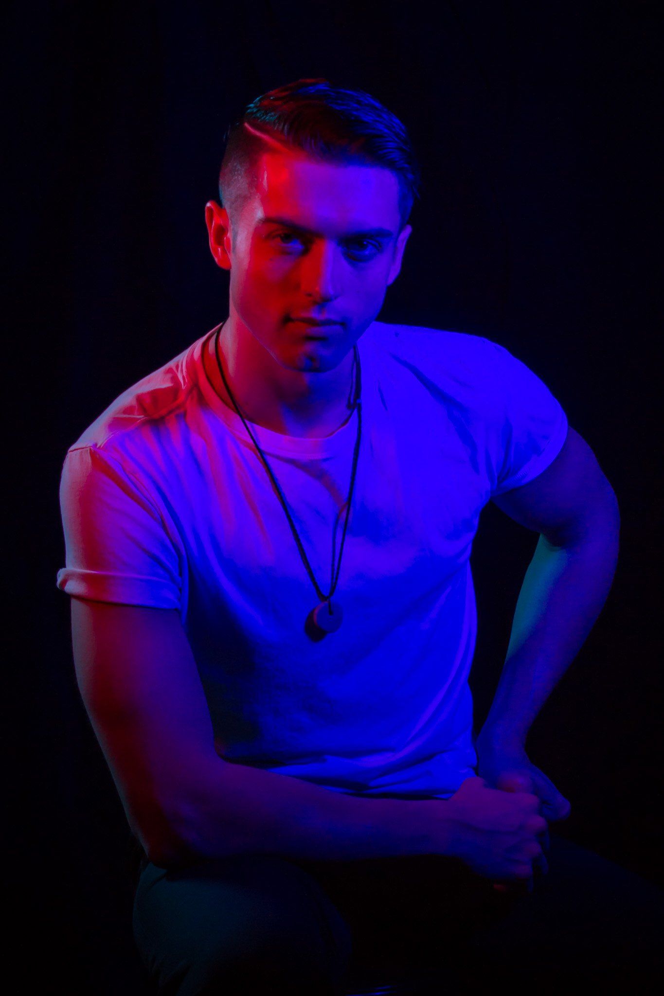 A man with short dark hair and white t-shirt lit with red light on one side and blue lighting on the other side.