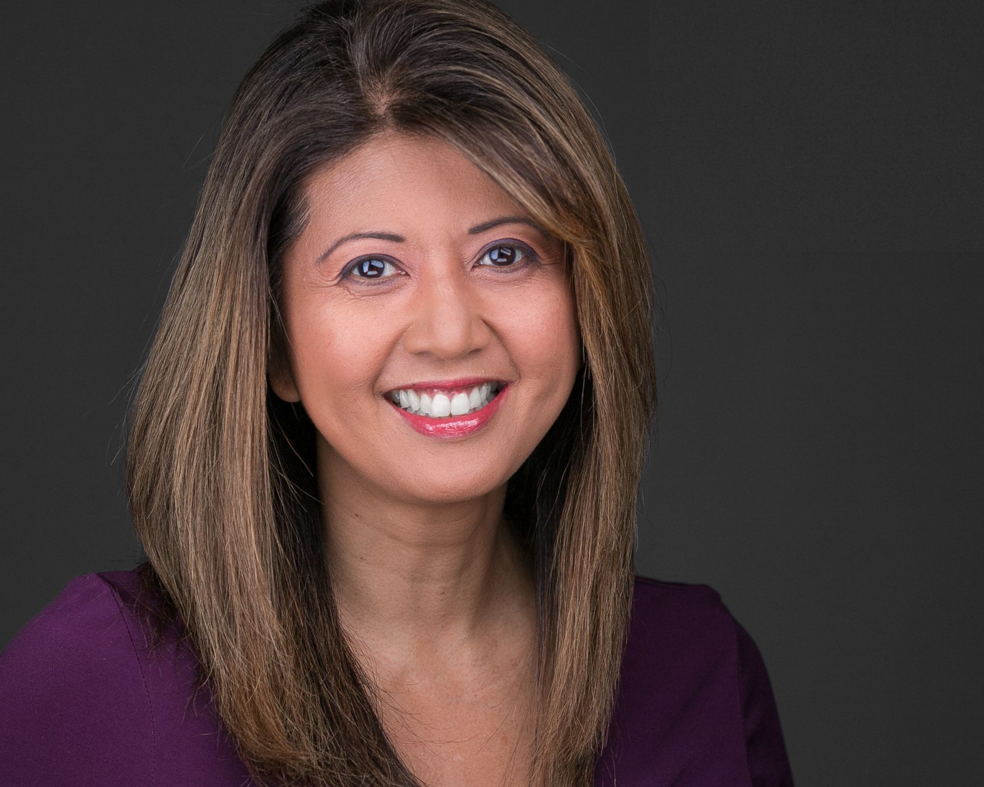 A woman with straight shoulder length brown hair in a purple shirt against a dark gray background.