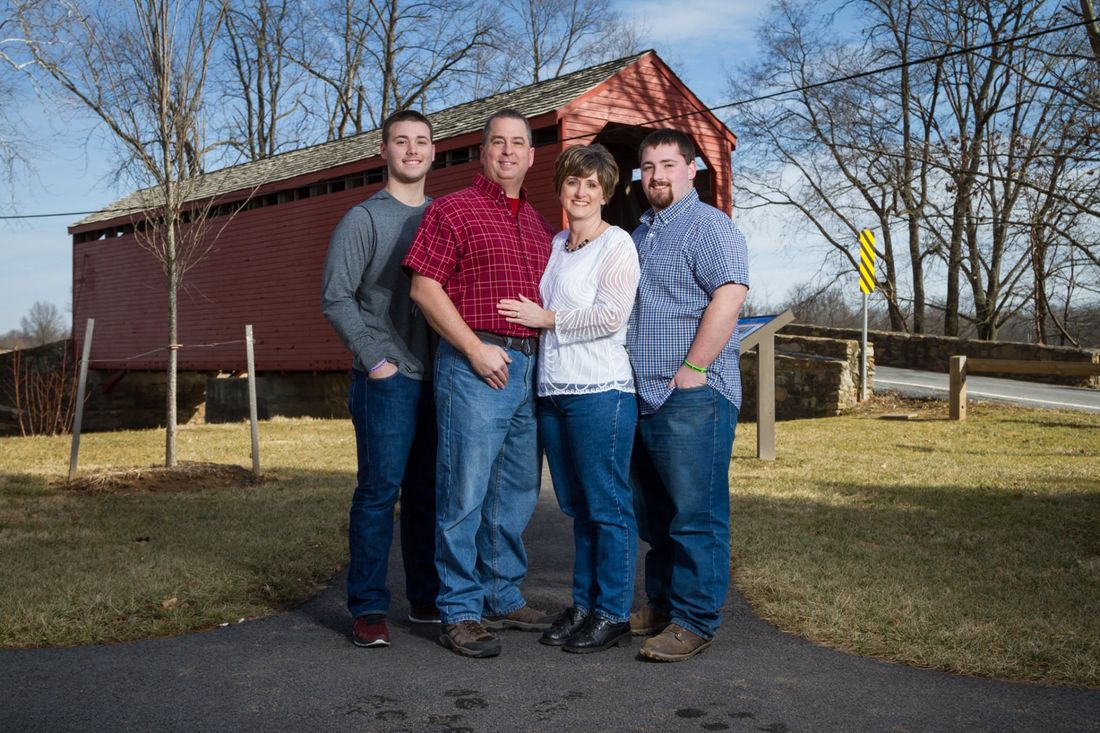 An outdoor portrait of a family of four, a man, woman and two sons standing in front of a rustic red covered bridge