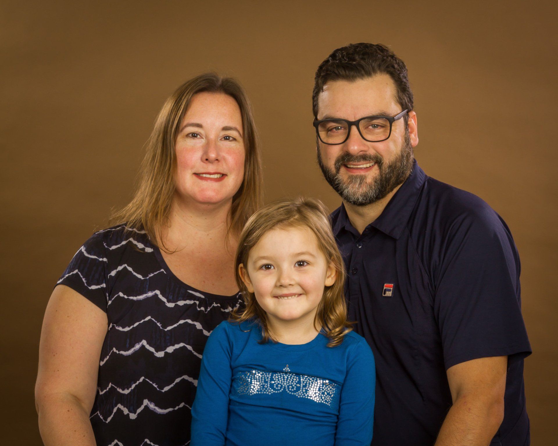 A couple and their young daughter posing in the studio for a portrait against a brown background.