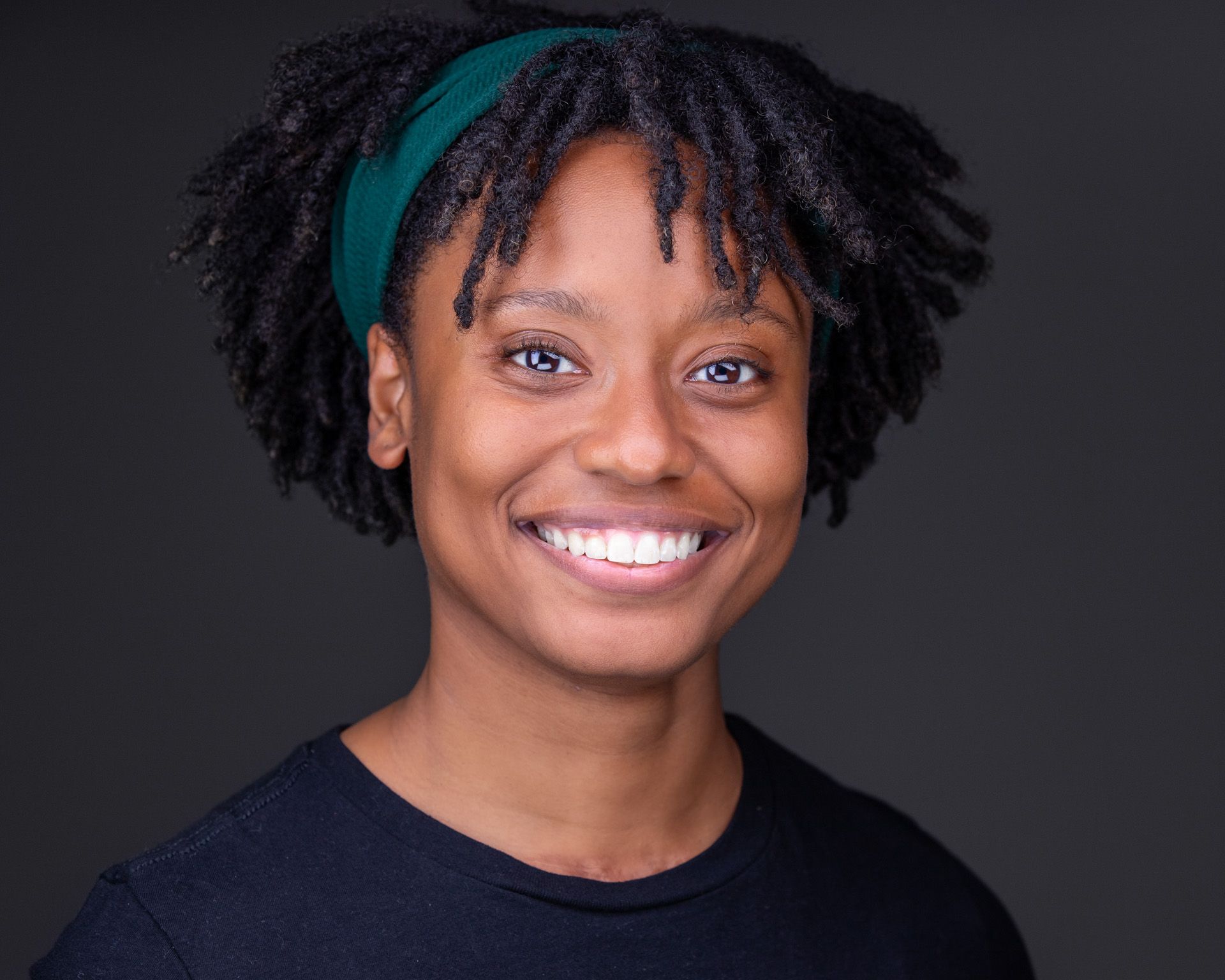 An African American woman with black braided hair and blue crew neck shirt against a dark gray background.