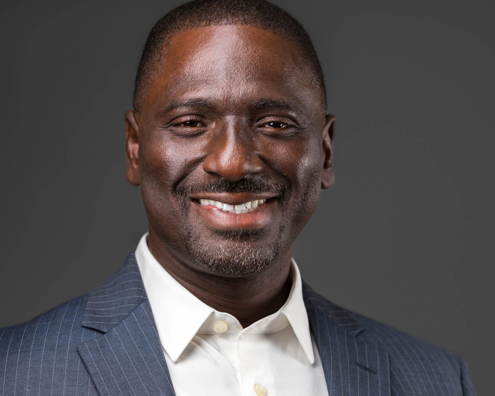 A headshot image of an African American man in a blue striped suit and white shirt against a gray background