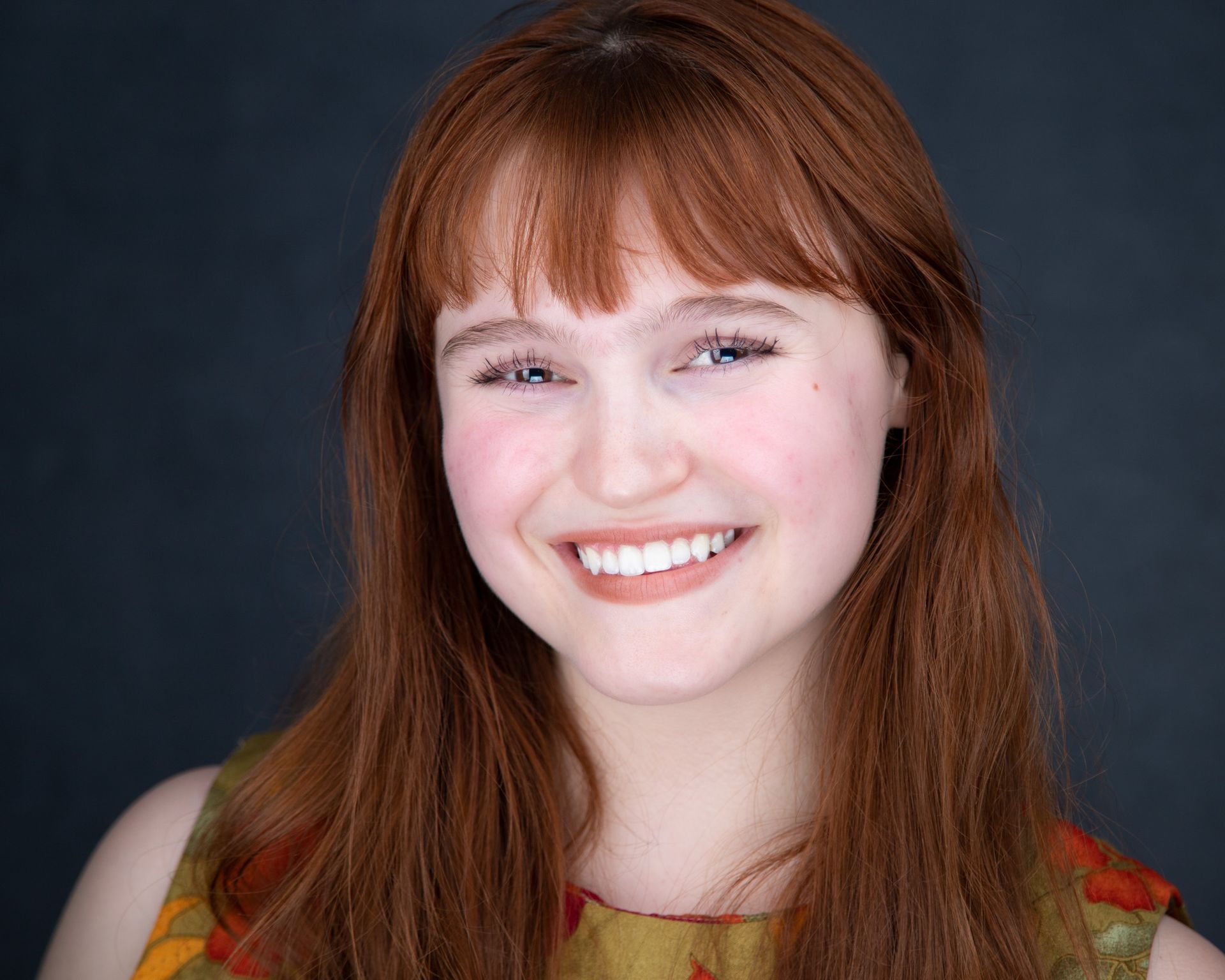 A headshot of a woman red strait hair wearing a red flowered top against a gray background.